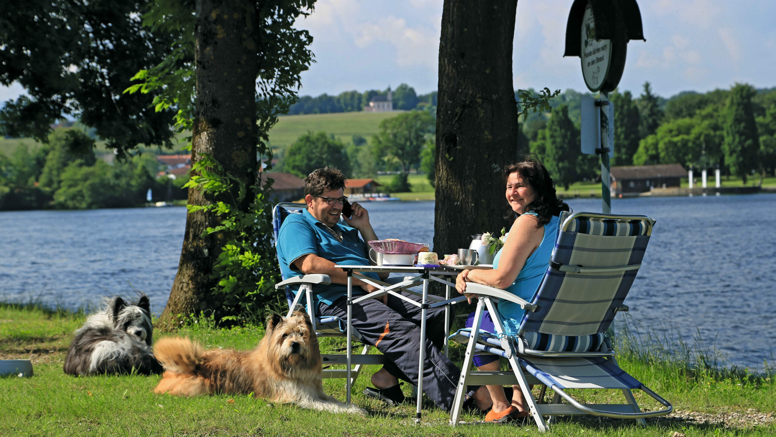 Zwei Personen an einem Campingtisch und zwei flauschige Hunde sitzen vor dem See