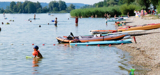Einige badende Leute und kleine Boote am Kiesstrand des Waginger Sees