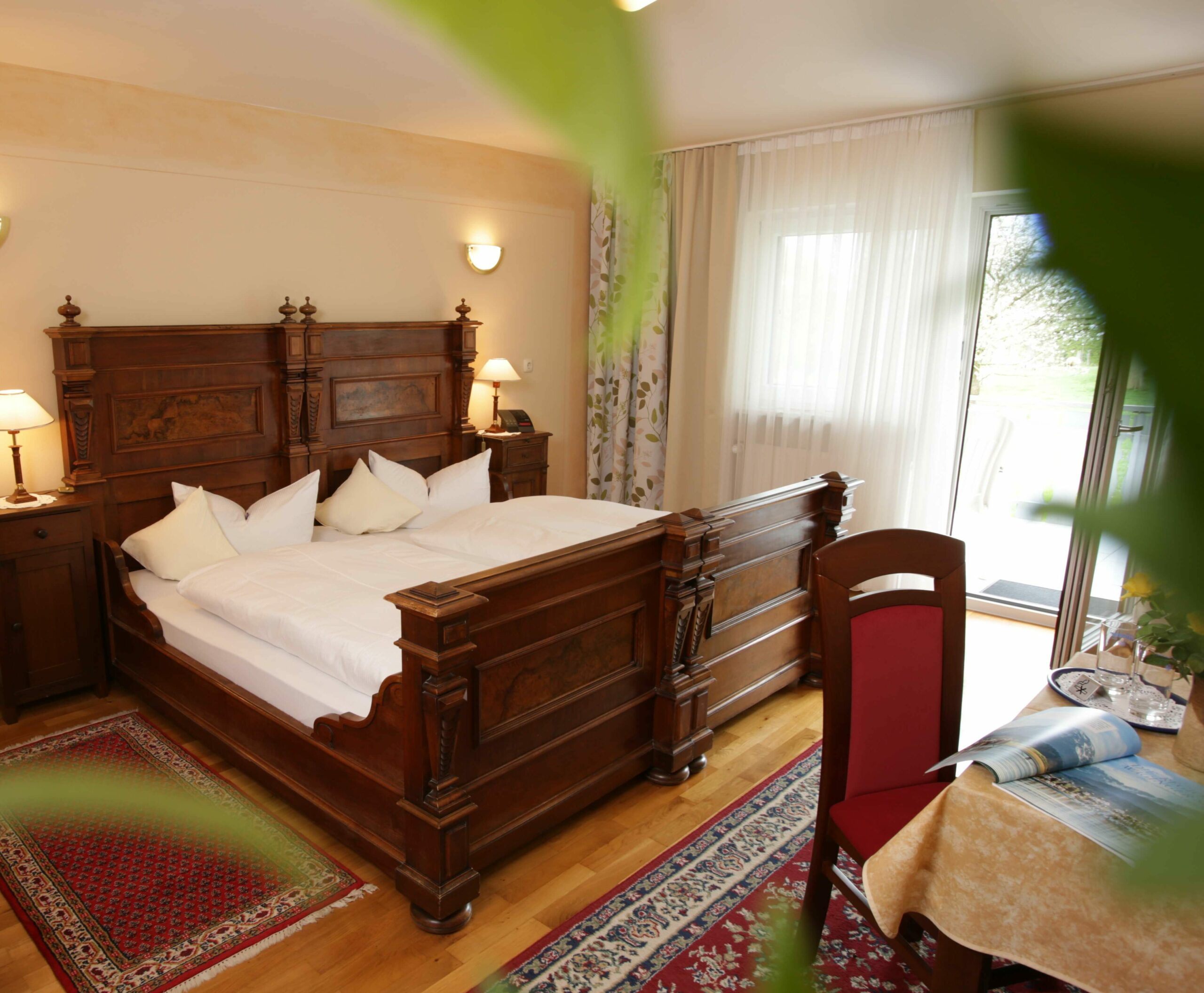 Ein frisch bezogenes großes Holzbett in einem sonnengefluteten Zimmer, vorne ein Tisch mit Stuhl und hinter dem Bett eine große Fensterfront und eine geöffnete Balkontüre