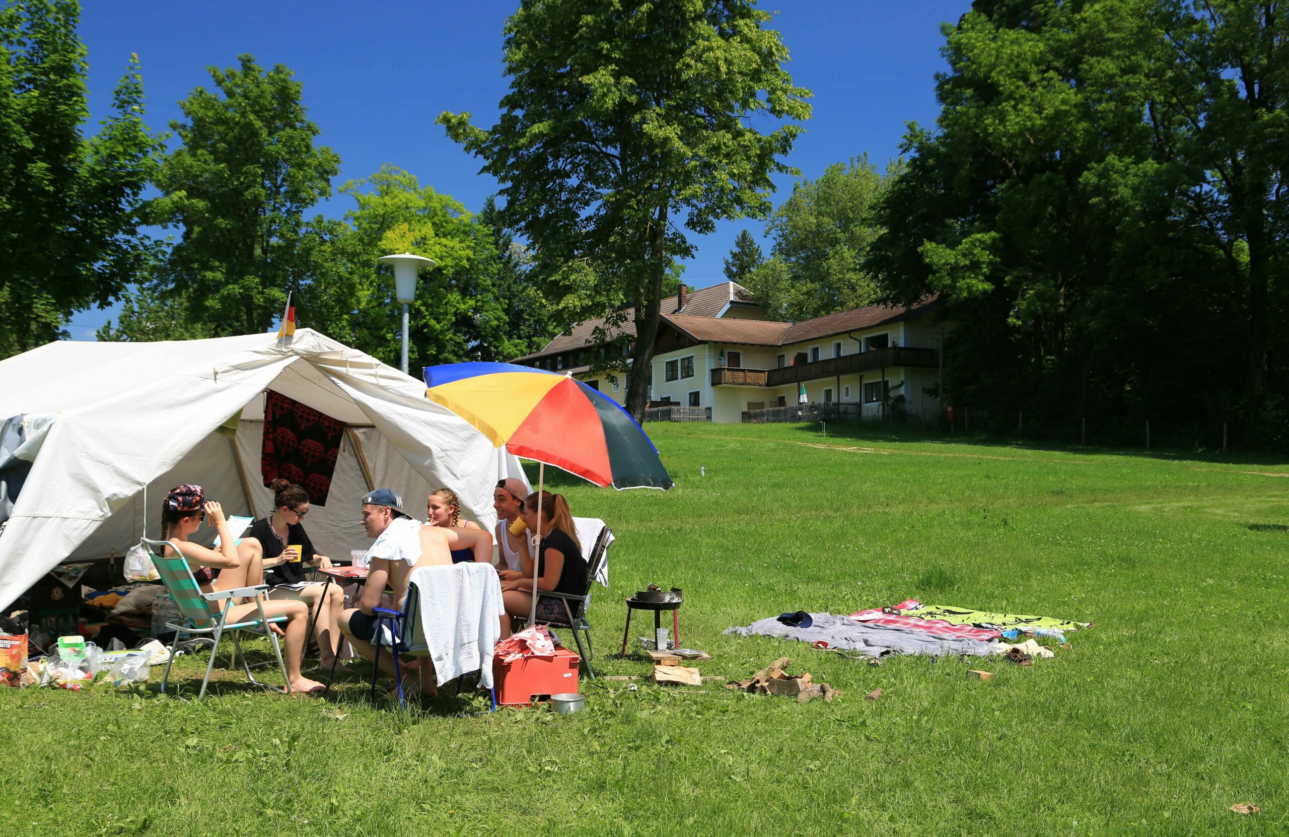 Eine Gruppe junger Leute sitzt mit einem kleinen Grill und Getränken im Sonnenschein vor einem Zelt auf der Campingwiese vor dem Hauptgebäude