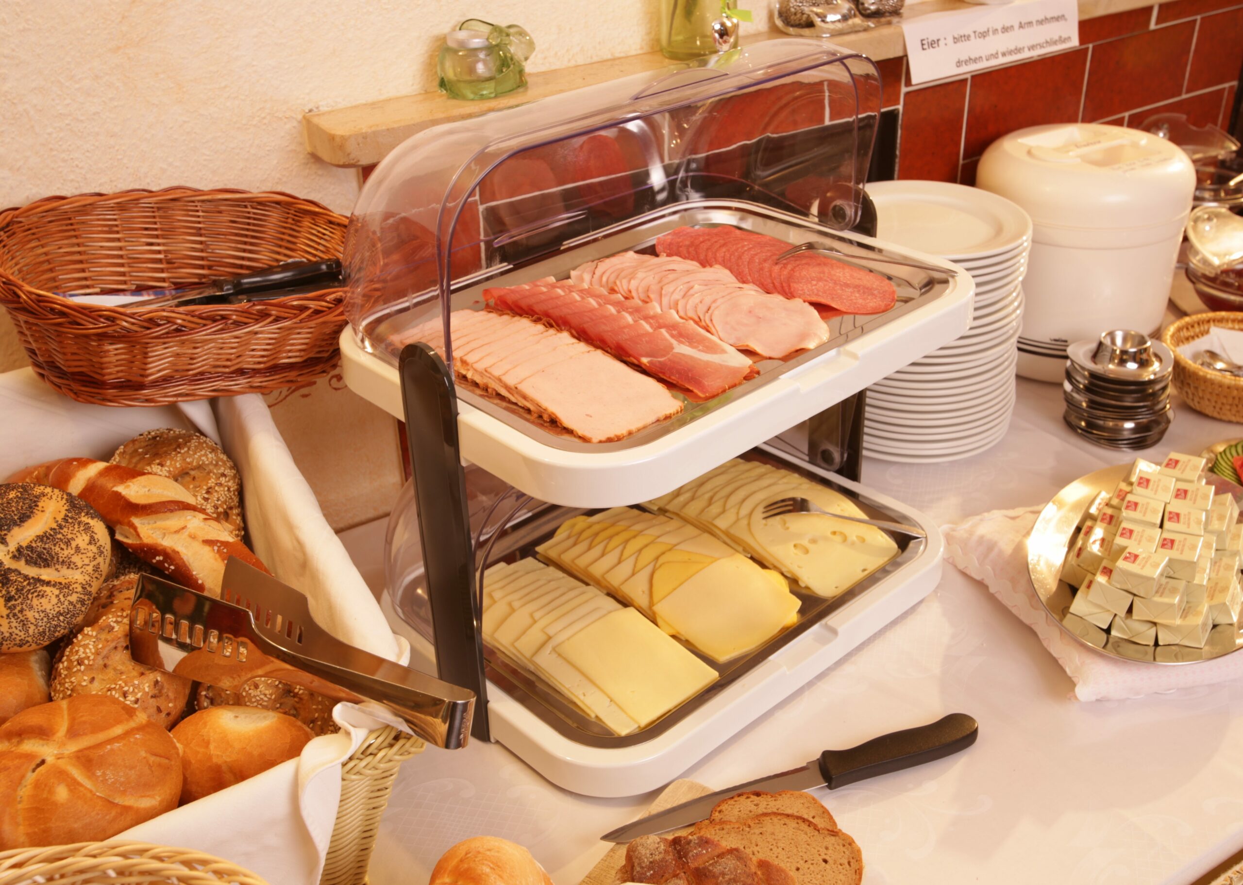 Eine Auswahl an Fleisch und Käse auf einem Buffet-Tisch. Daneben frische Semmeln und Laugenstangen in einem Korb und Butter, sowie gestapelte Teller
