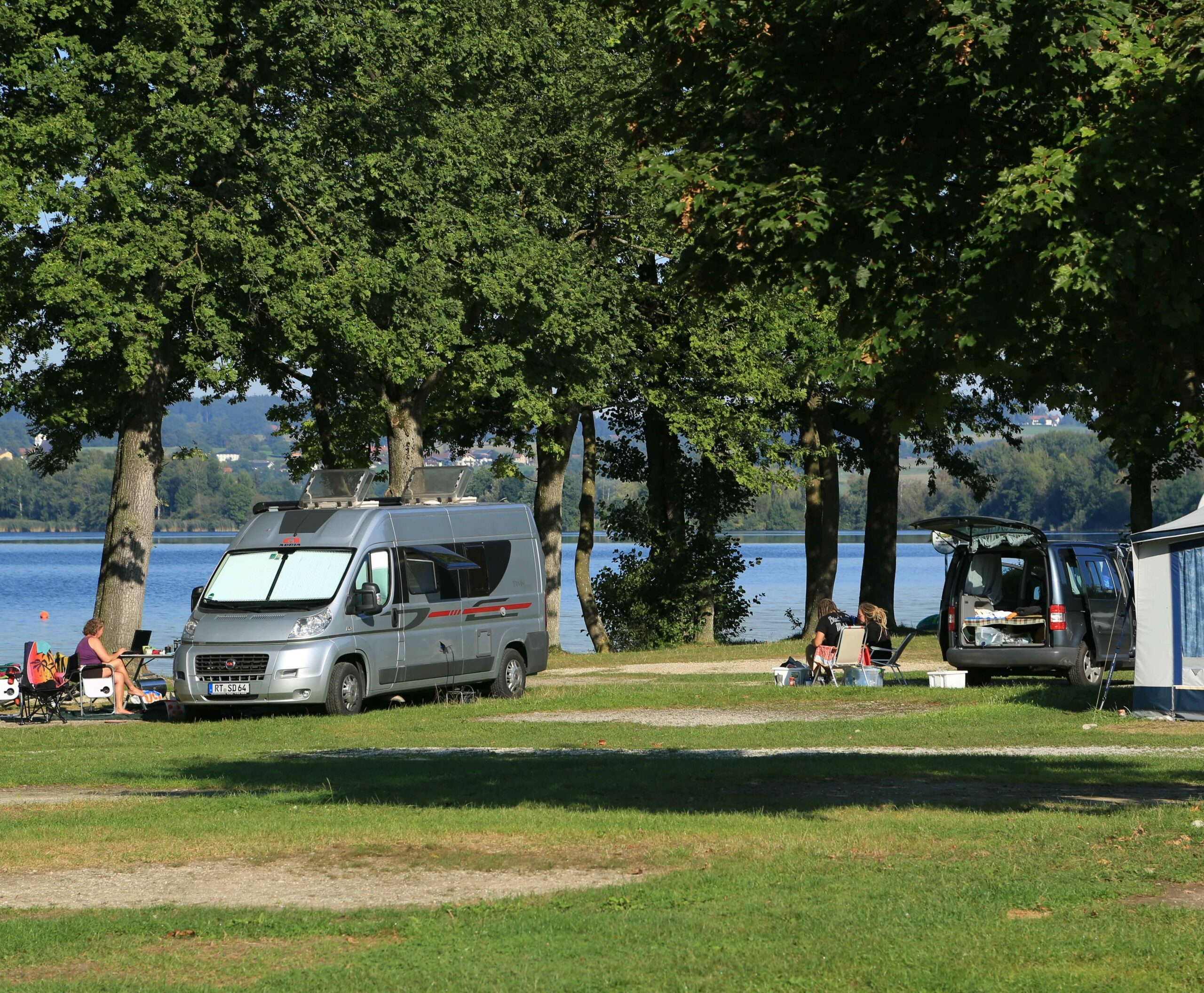 Mehrere Camper und Campingbusse auf der Campingwiese vor dem See.