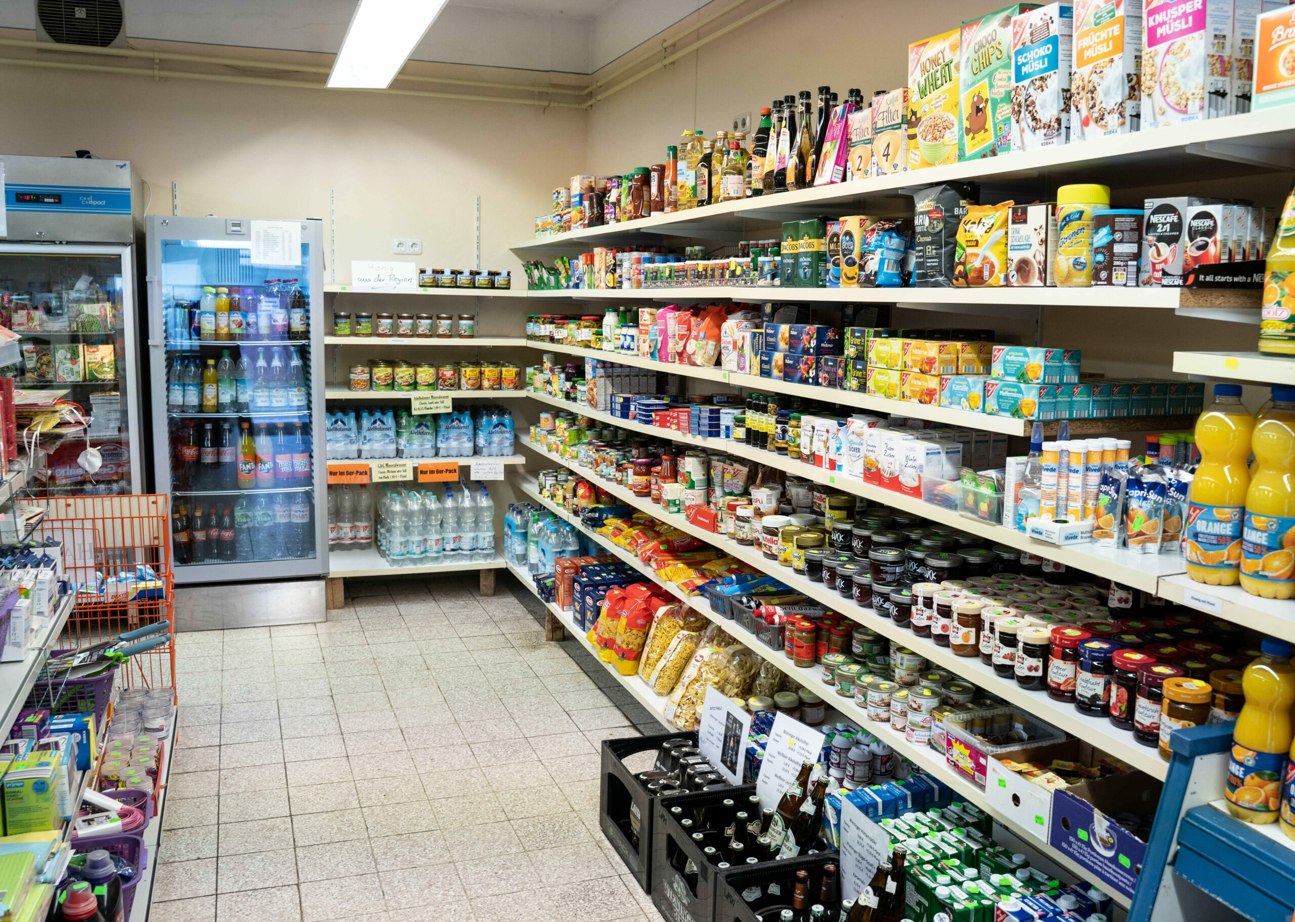 Mehrere voll befüllte Regale eines Supermarkts, links ein Kühlschrank mit Getränken