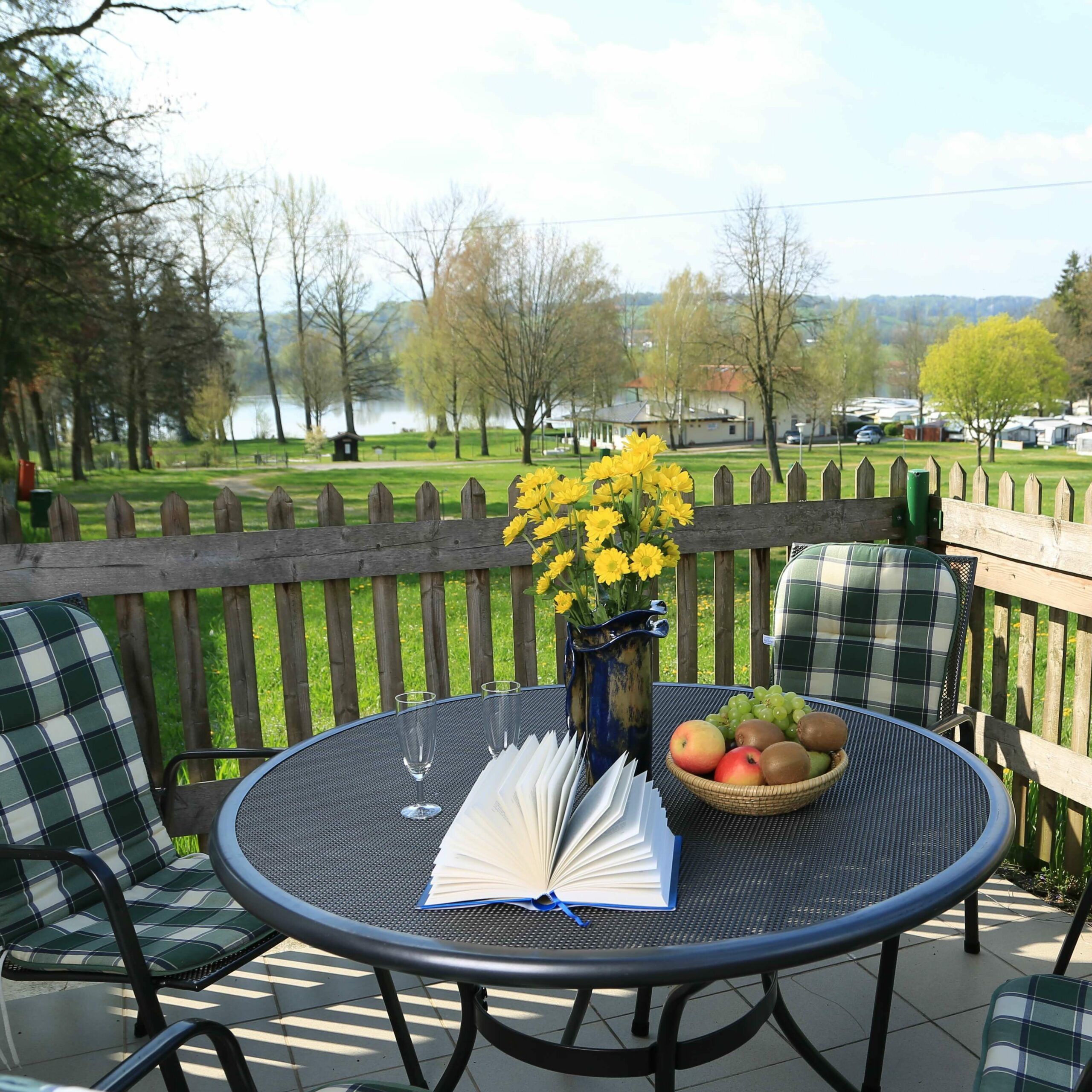 Eine eingezäunte Terrasse einer Ferienwohnung mit einem großen runden Tisch und mehreren Stühlen, im Hintergrund eine saftige Wiese und der Waginger See