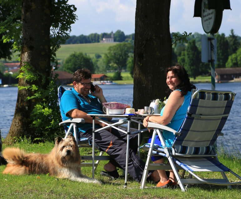 Zwei Personen an einem Campingtisch und zwei flauschige Hunde sitzen vor dem See