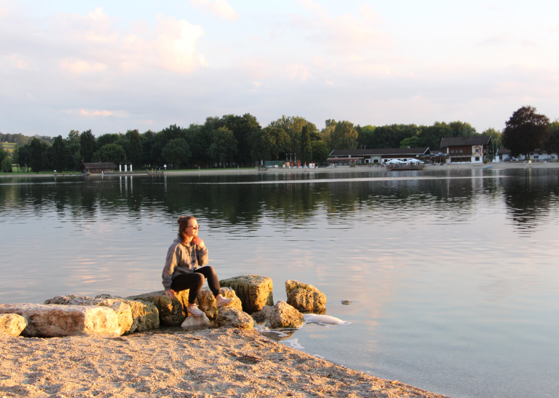 Eine Frau sitzt auf größeren Steinen am Ufer des Waginger Sees bei Sonnenuntergang