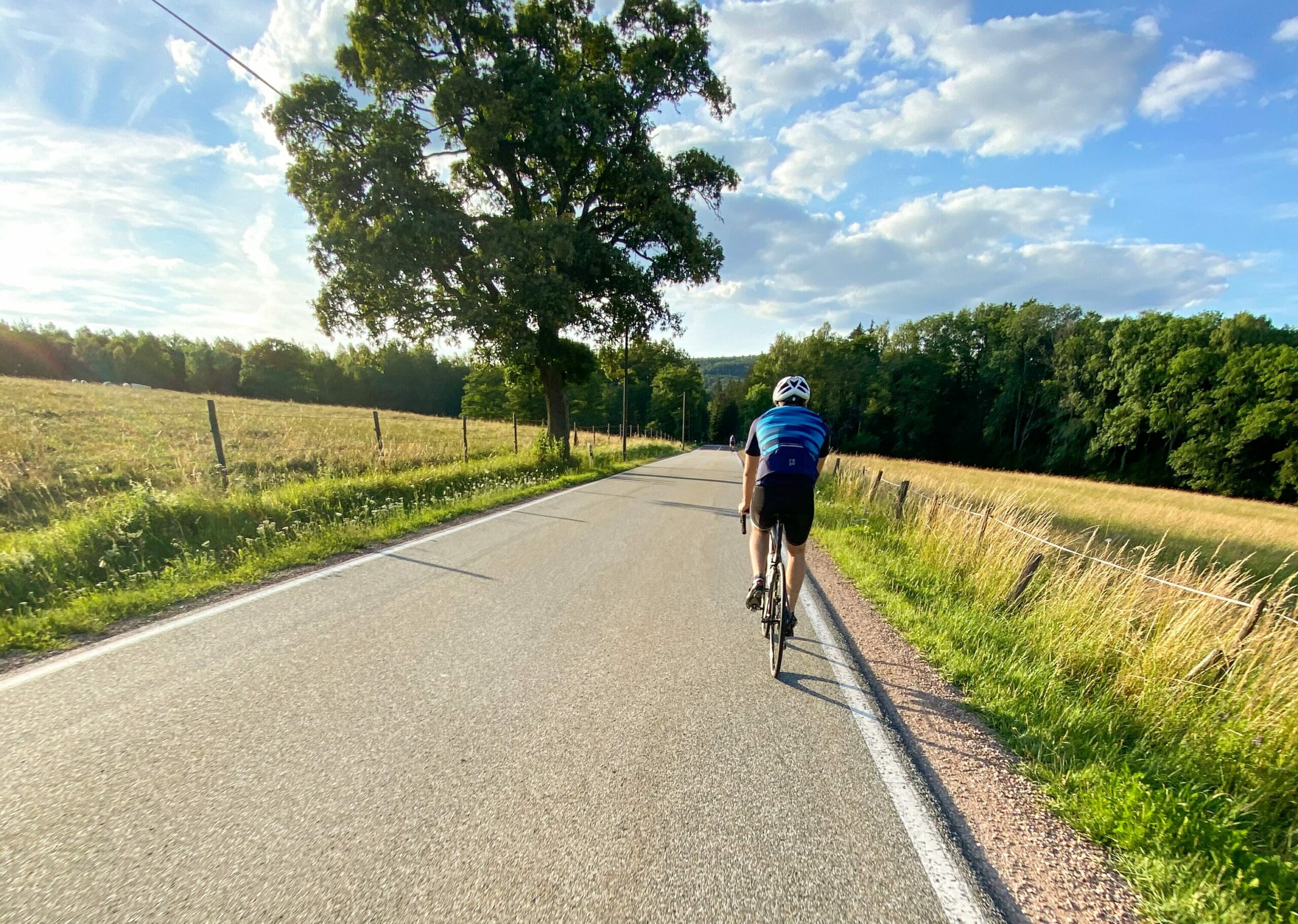 Zwei Rennradfahrer mit Helm und Sportkleidung fährt auf einer sonnigen Straße zwischen zwei Feldern