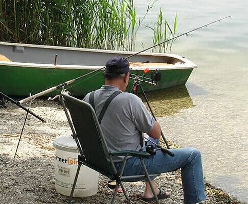 Ein Angler sitzt mit Angelausrüstung und einem kleinen Boot am Ufer des Sees